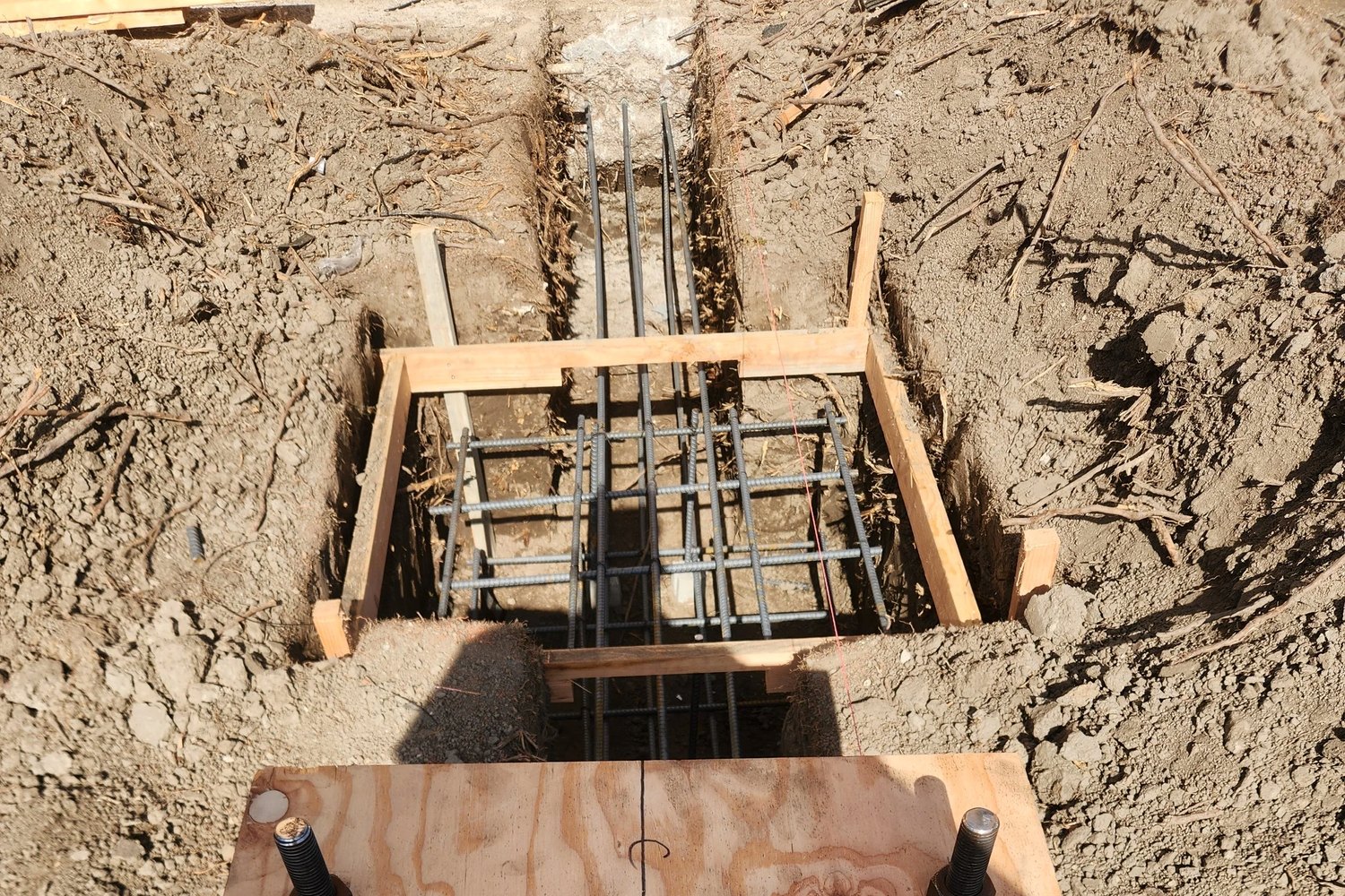 A footing trench with its rebar cage and form boards, hold-down anchors set on plywood