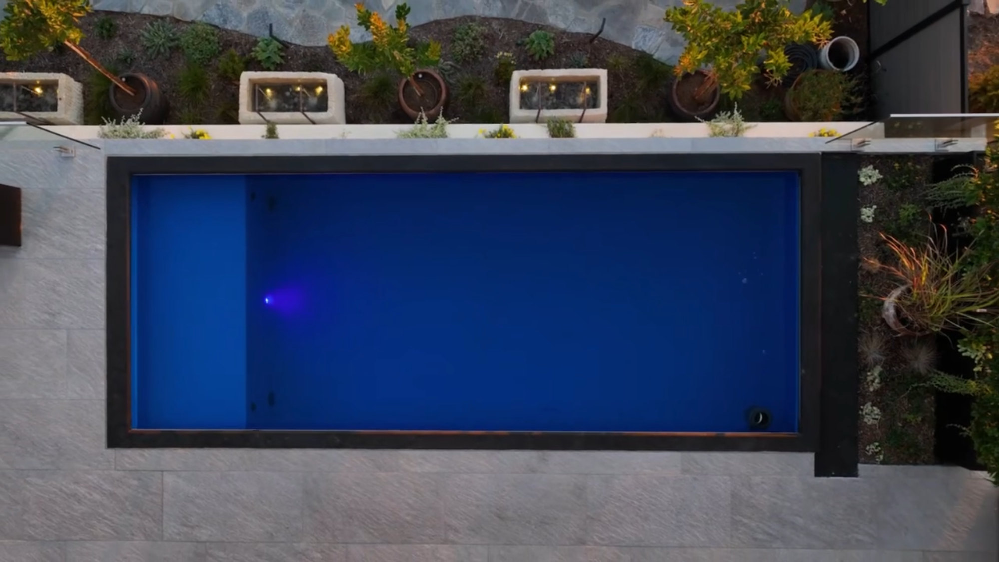 The Modpool container pool lit at night, seen from above, with the planted terrace and the lit niches behind it