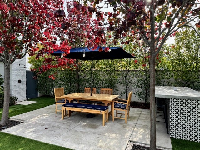 The rebuilt back yard, a teak dining table under flowering trees beside the patterned tile of the outdoor counter.